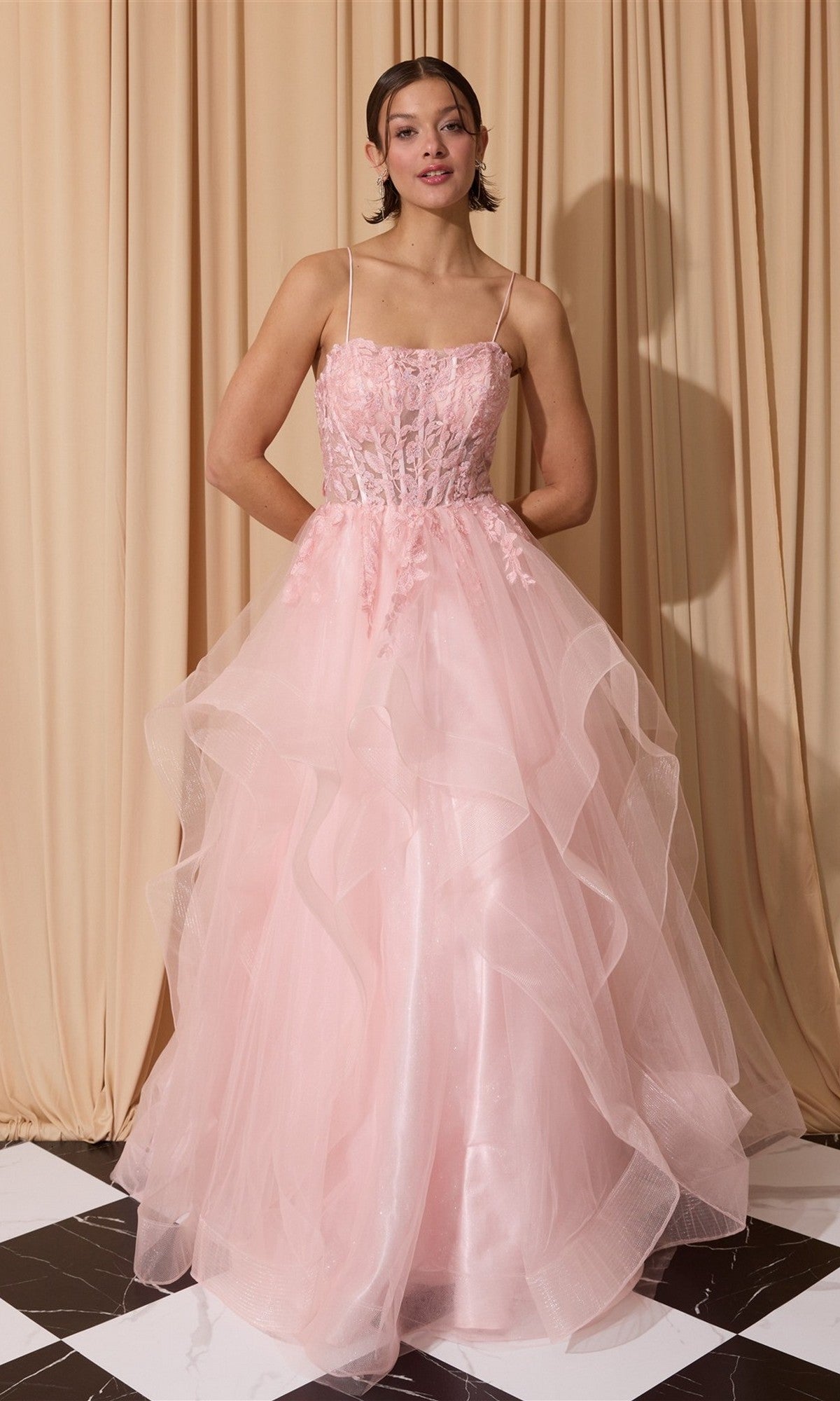 A woman stands on a black and white tiled floor in the Eureka EK167 long pink prom dress with a layered tulle A-line skirt and delicate floral details, set against a beige curtain backdrop.